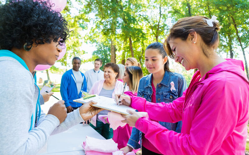Diverse people registering for charity breast cancer awareness race Diverse group of adults and teenagers are lined up at registration table in sunny park. They are signing up to run in charity 5k race or marathon to raise money for breast cancer research. People are wearing pink athletic clothing and breast cancer awareness ribbons.